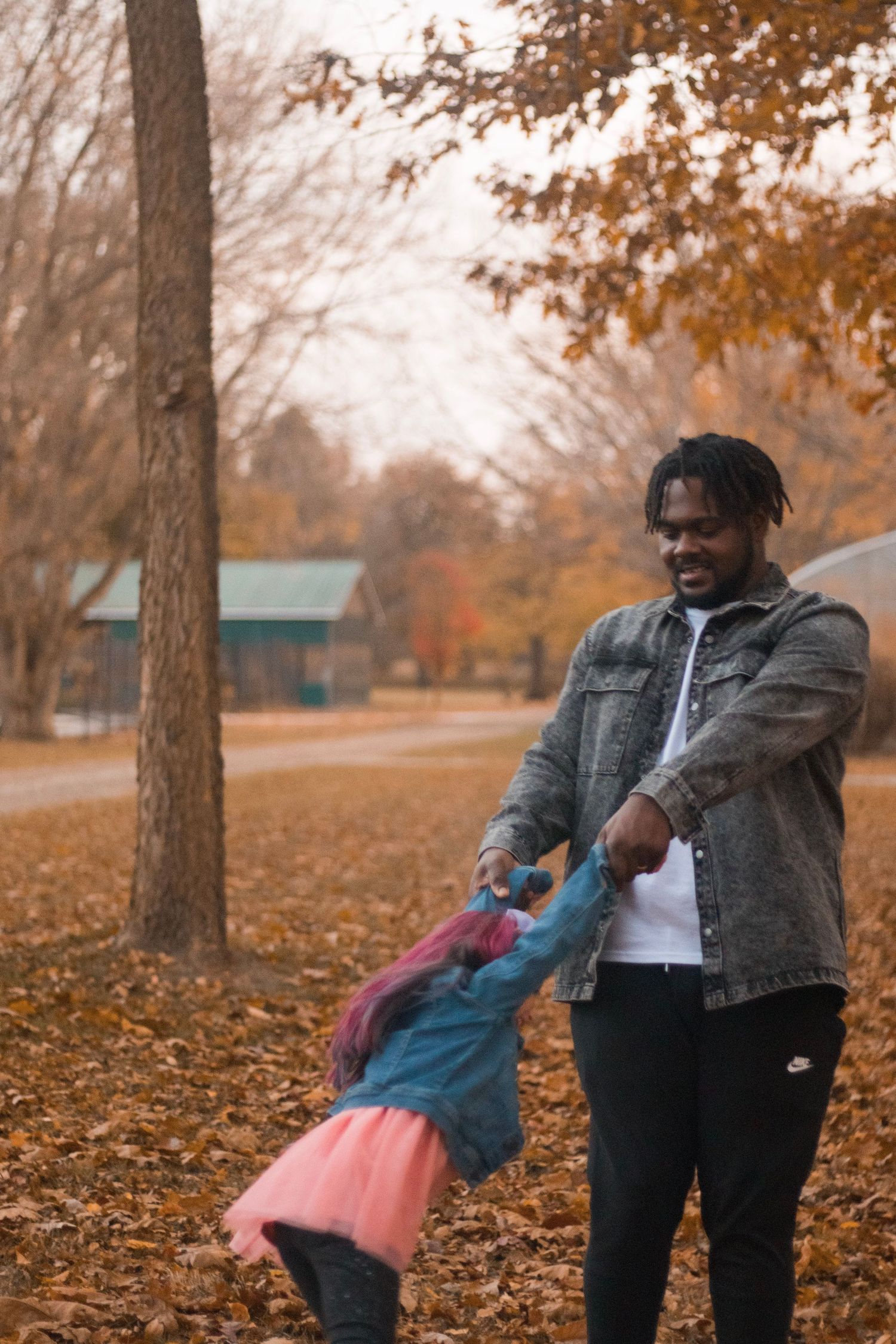 Father playfully swinging young daughter with pink hair in colorful autumn park.