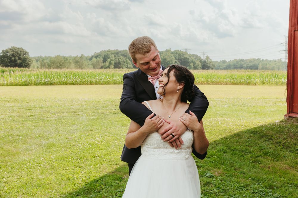Groom embraces bride from behind as they laugh together in front of a red barn and green cornfield.