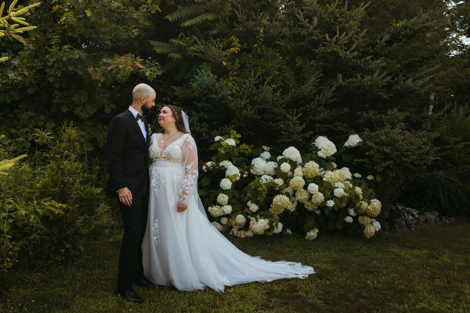 Bride and groom share a romantic moment in a lush garden with white hydrangeas, bride in elegant lace gown.