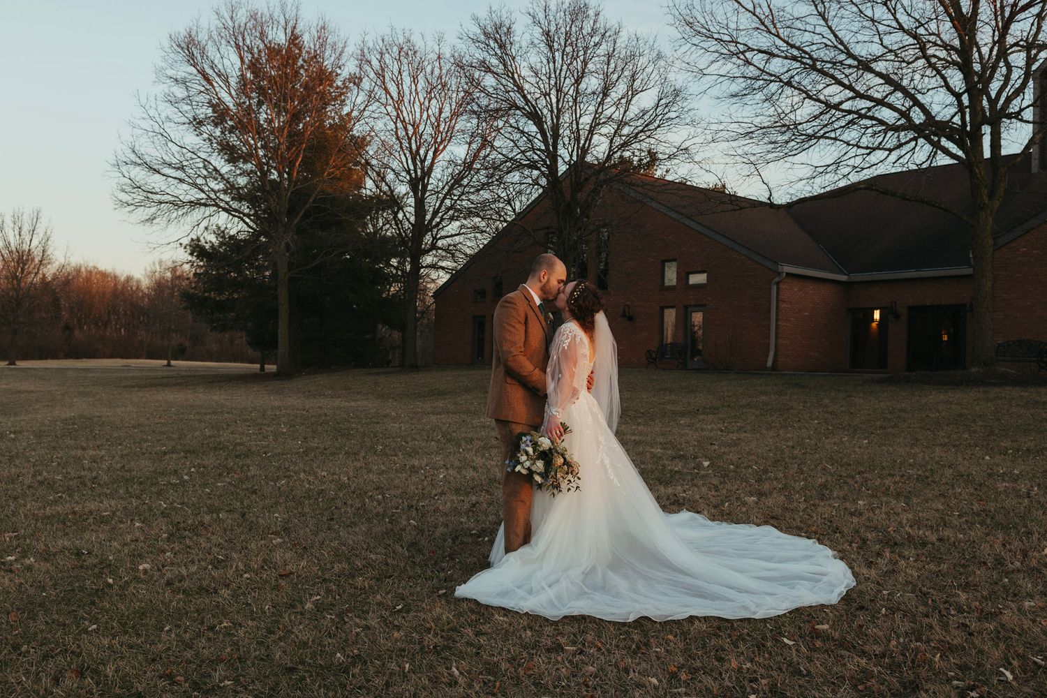 Bride and groom sharing a romantic sunset kiss in an open field with autumn trees and brick building backdrop.