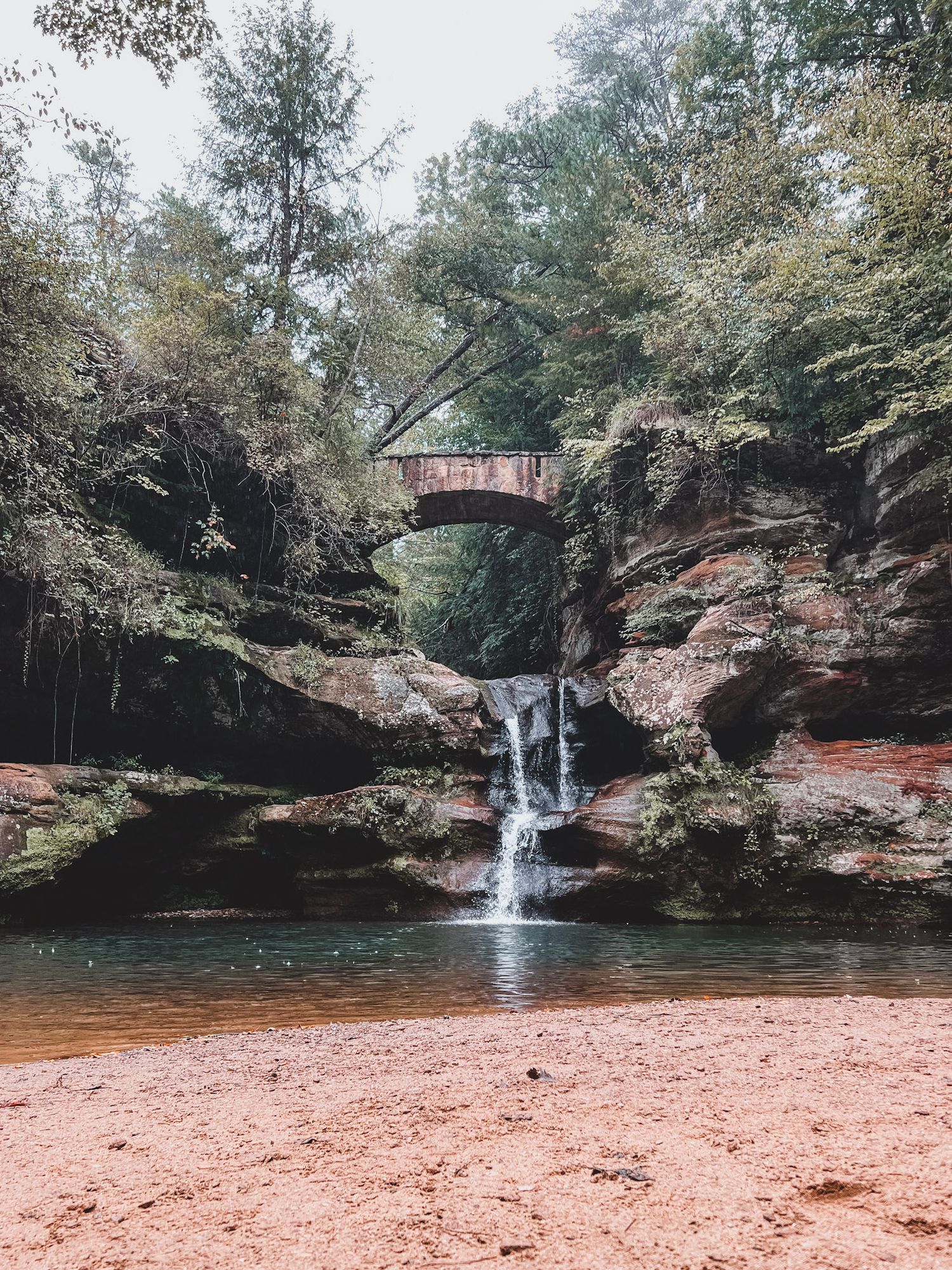 Forest waterfall beneath a stone bridge surrounded by lush green trees and rocky cliffs, Hocking Hills, Ohio