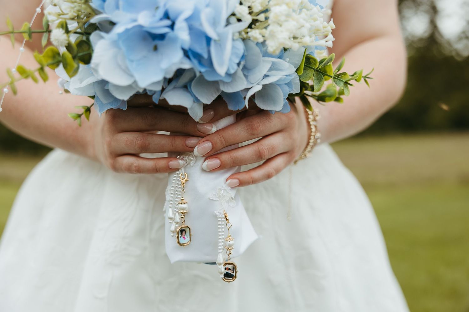 Bride holding blue hydrangea bouquet with white ribbon and gold rings, wearing white wedding dress with elegant manicured nails.