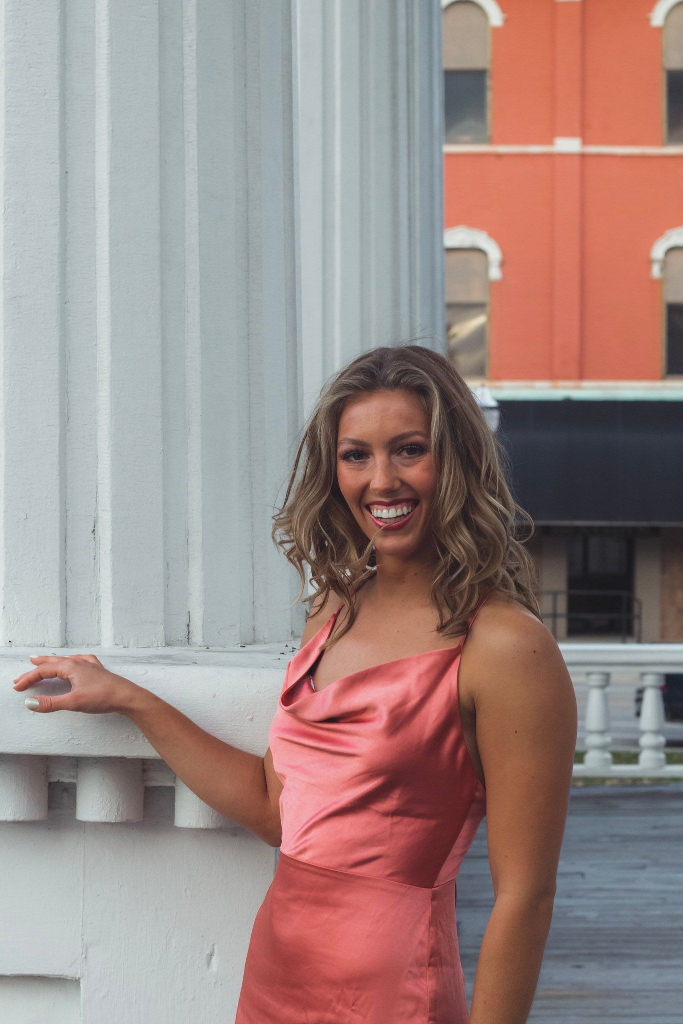 Smiling woman in pink satin dress leaning against white column outdoors.