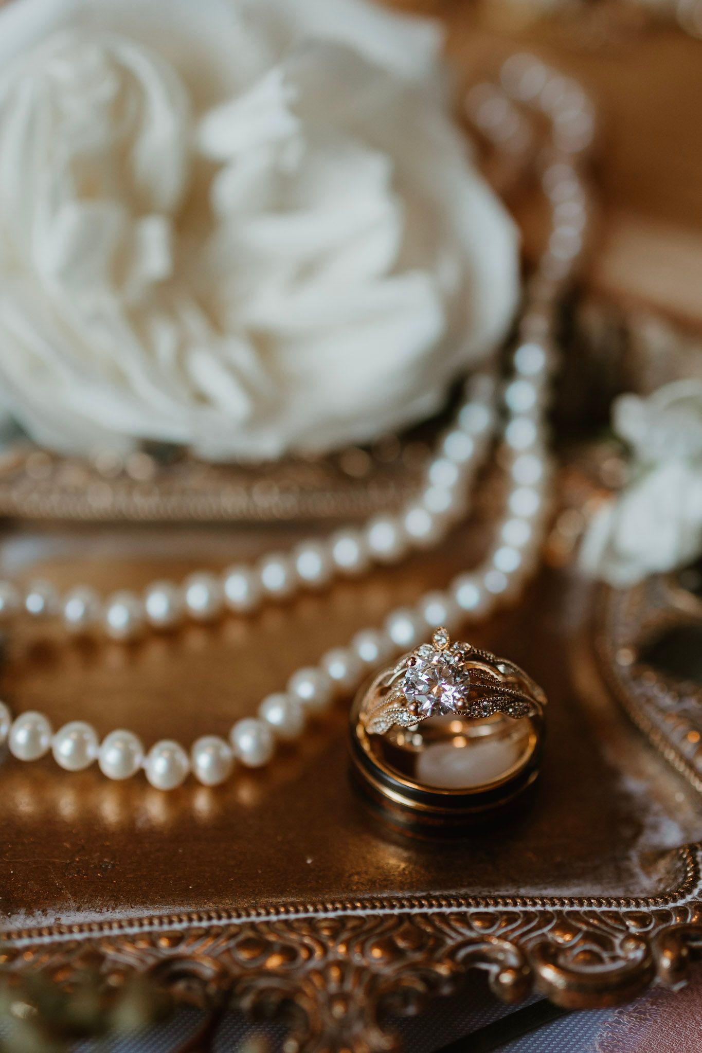 Gold wedding rings resting on pearl necklace atop ornate golden tray with white peony bouquet.