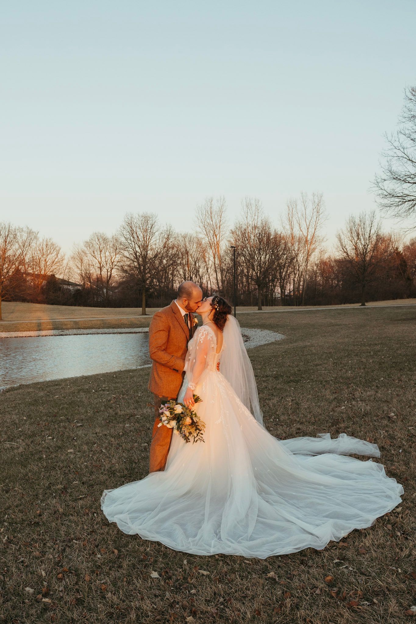 Bride and groom kissing at golden hour near pond, bride in long veil and groom in rust suit.
