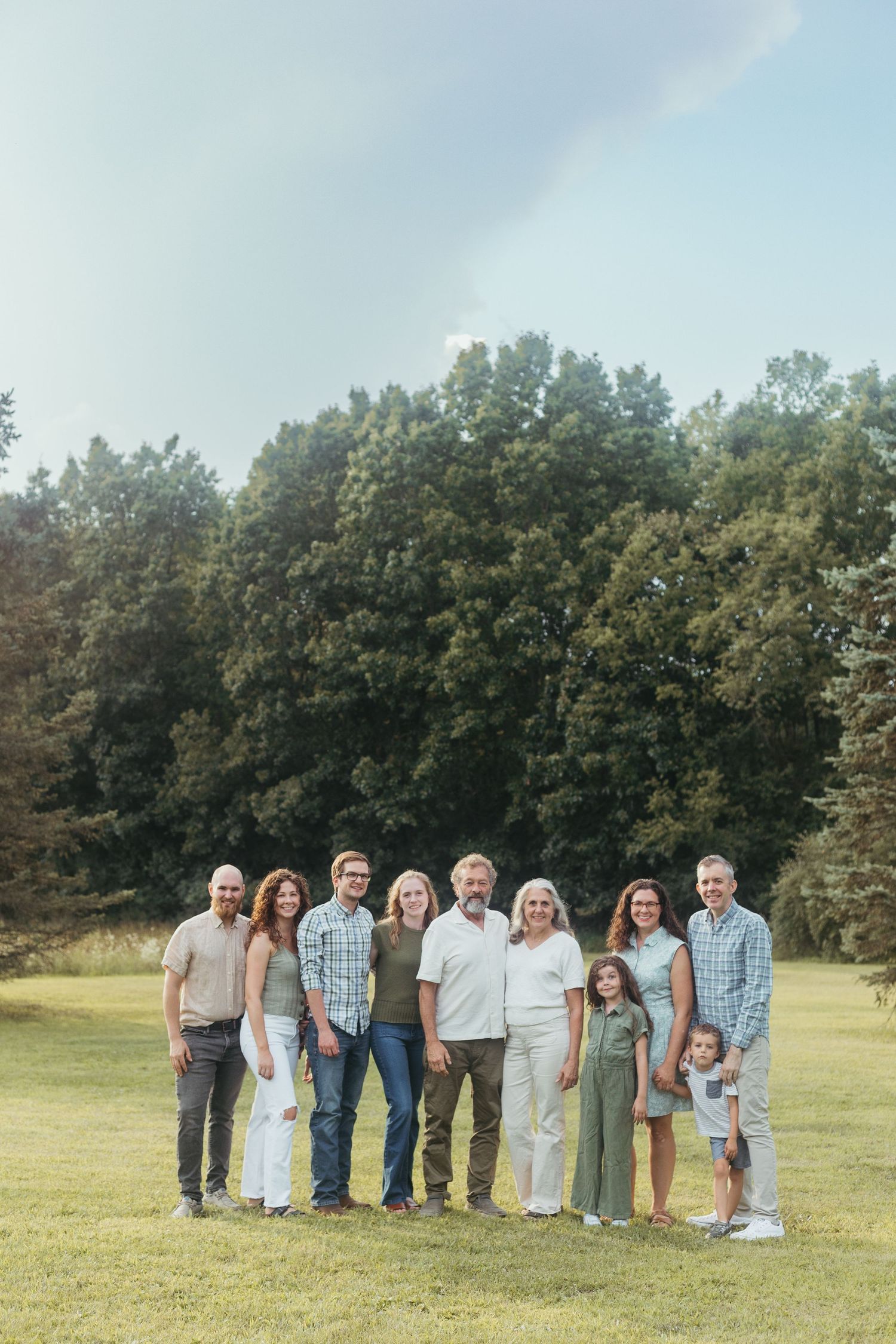 A multi-generational family poses together on a green lawn surrounded by tall trees on a cloudy day.