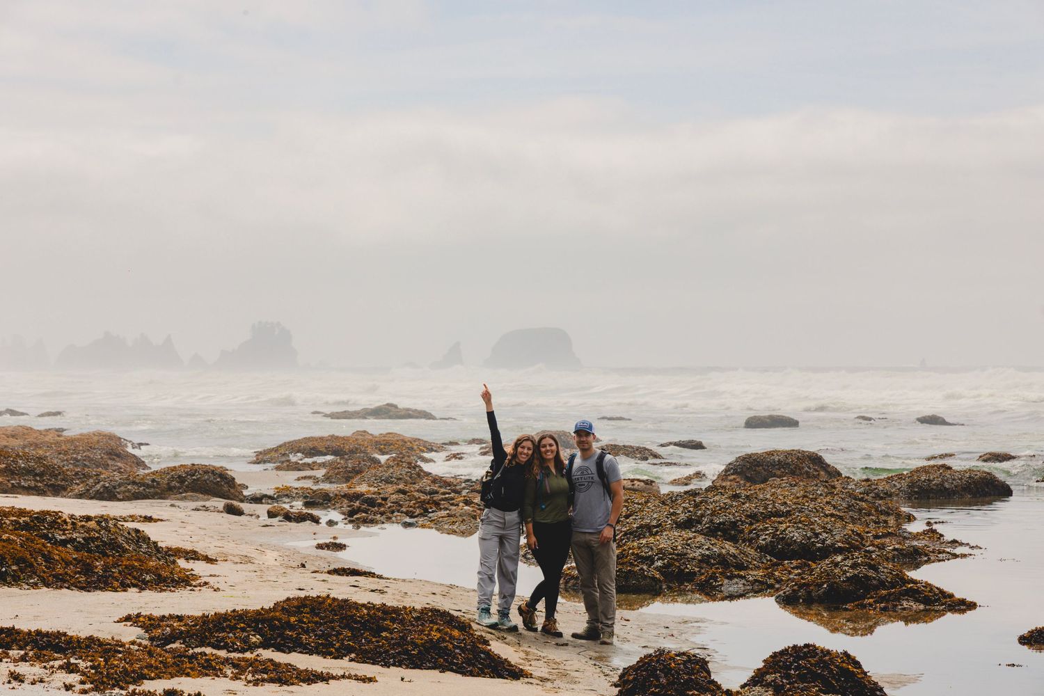 Group of three posing on a rugged Pacific coast beach with misty waves and rocky sea stacks in background.