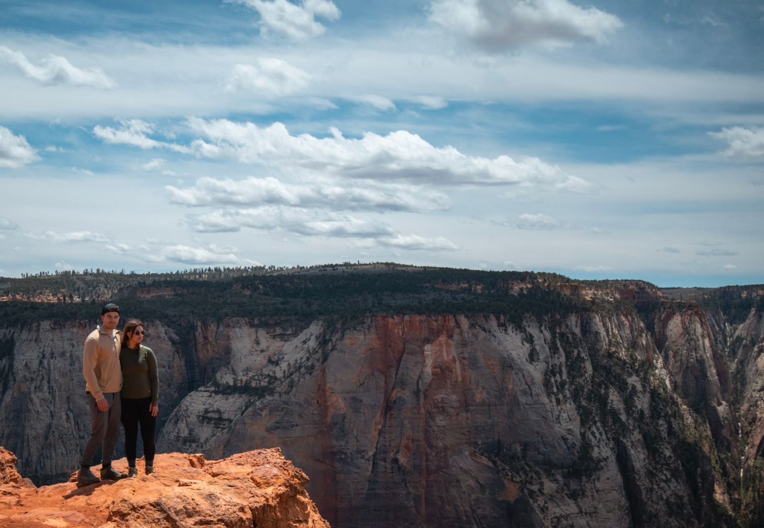 Couple standing at a dramatic canyon overlook with layered red rock cliffs and expansive desert views, Zion National Park