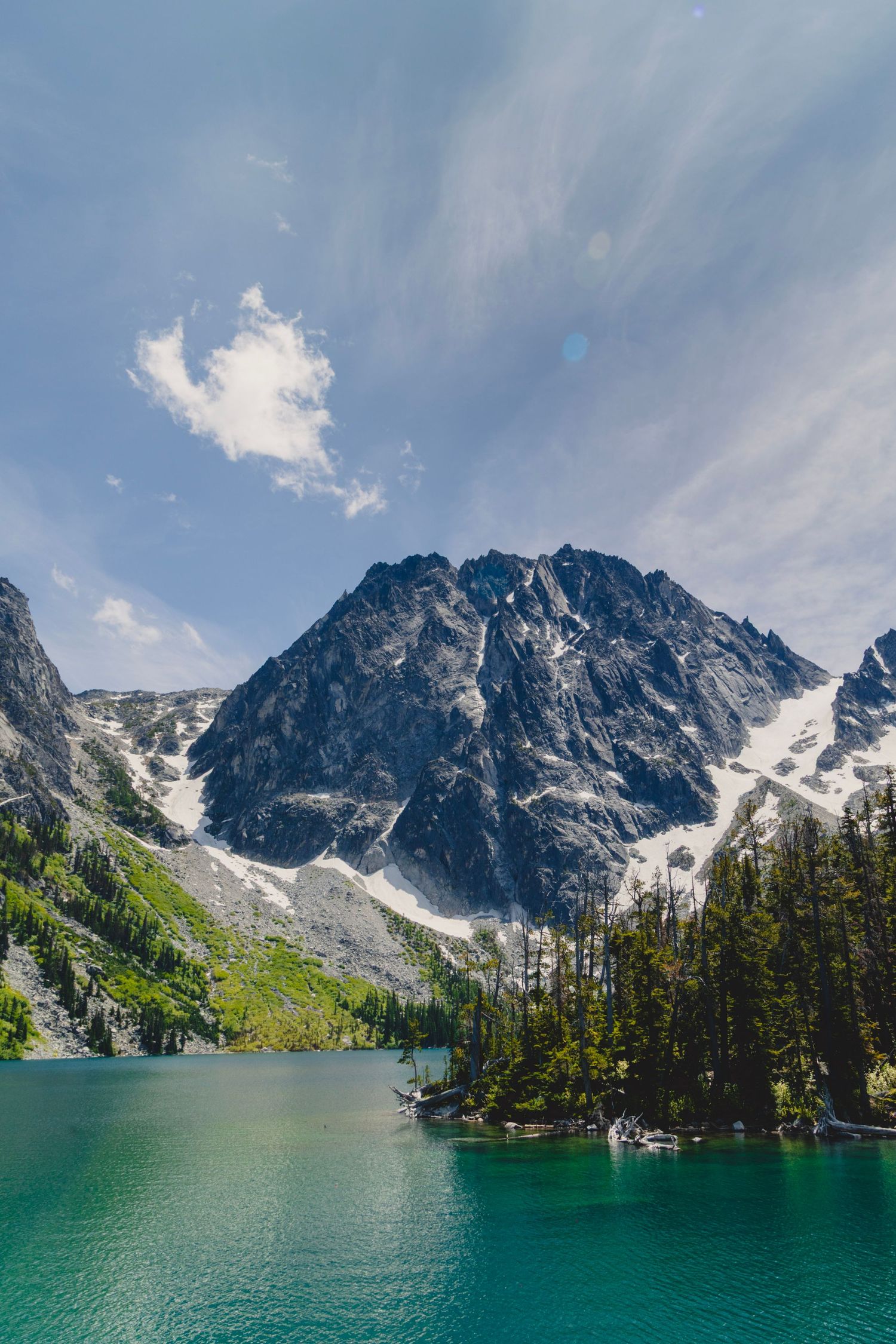 Turquoise alpine Colchuck Lake surrounded by rugged mountain peaks and evergreen trees under a blue summer sky
