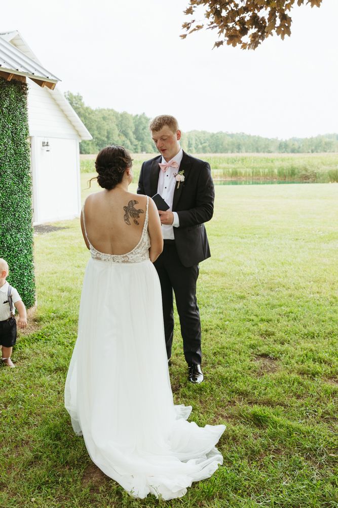 Groom reading vows to tattooed bride in backless white dress near lakeside ivy-covered building.