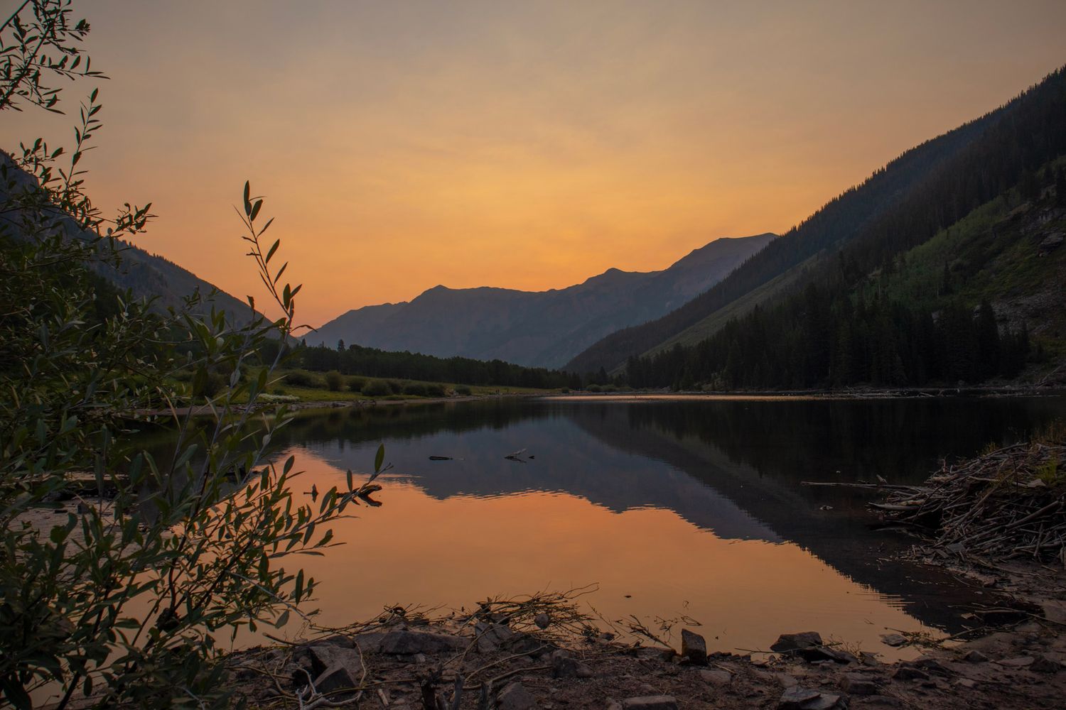 Maroon Bells at sunrise reflected in calm alpine lake with evergreen trees and dramatic Colorado mountain peaks