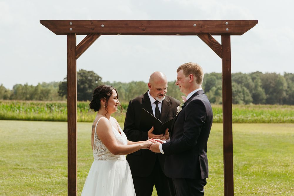 Outdoor August wedding ceremony in New Boston, Michigan with bride and groom exchanging vows under a wooden arbor