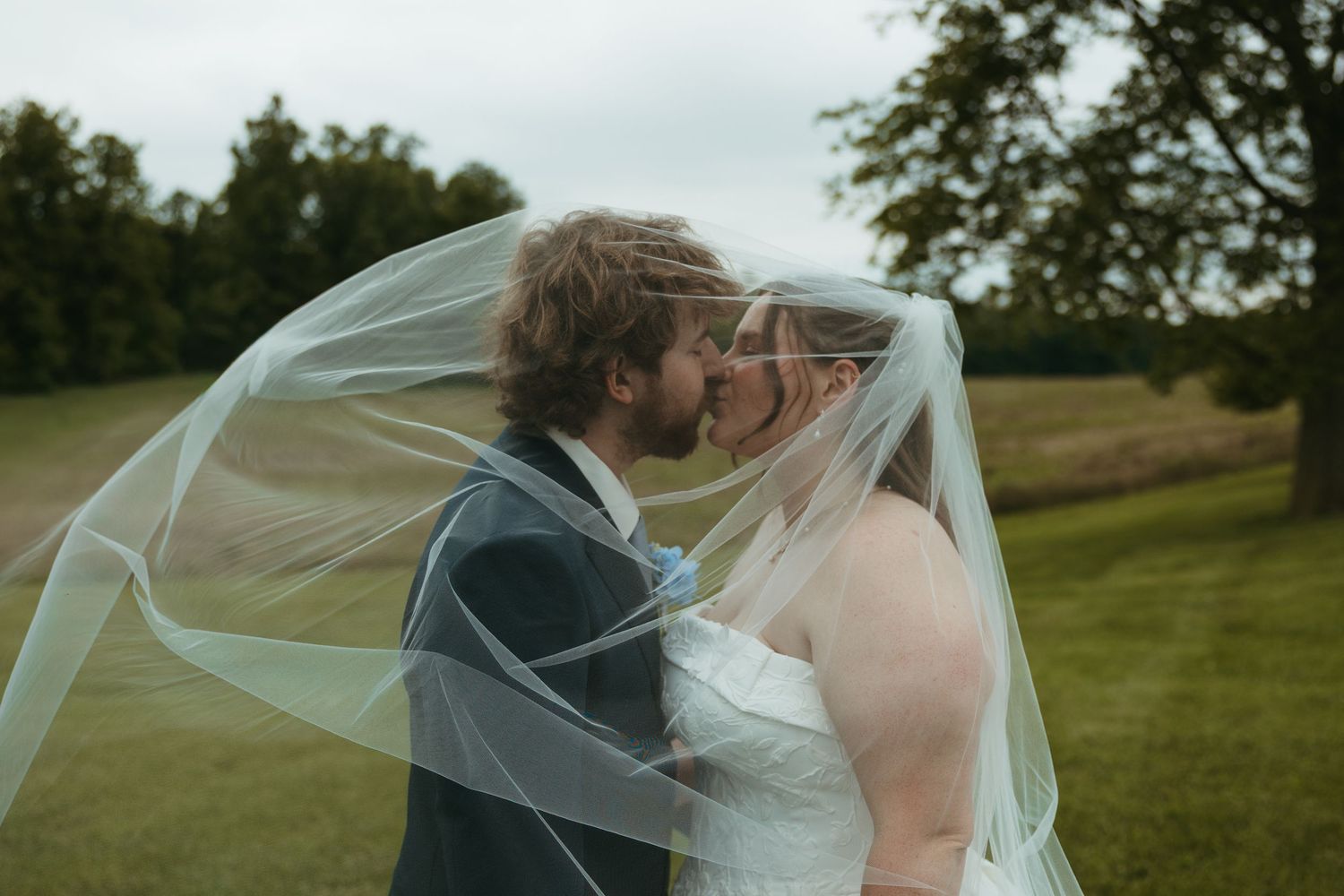 Bride and groom sharing a kiss under a flowing wedding veil outdoors on a lush green lawn.