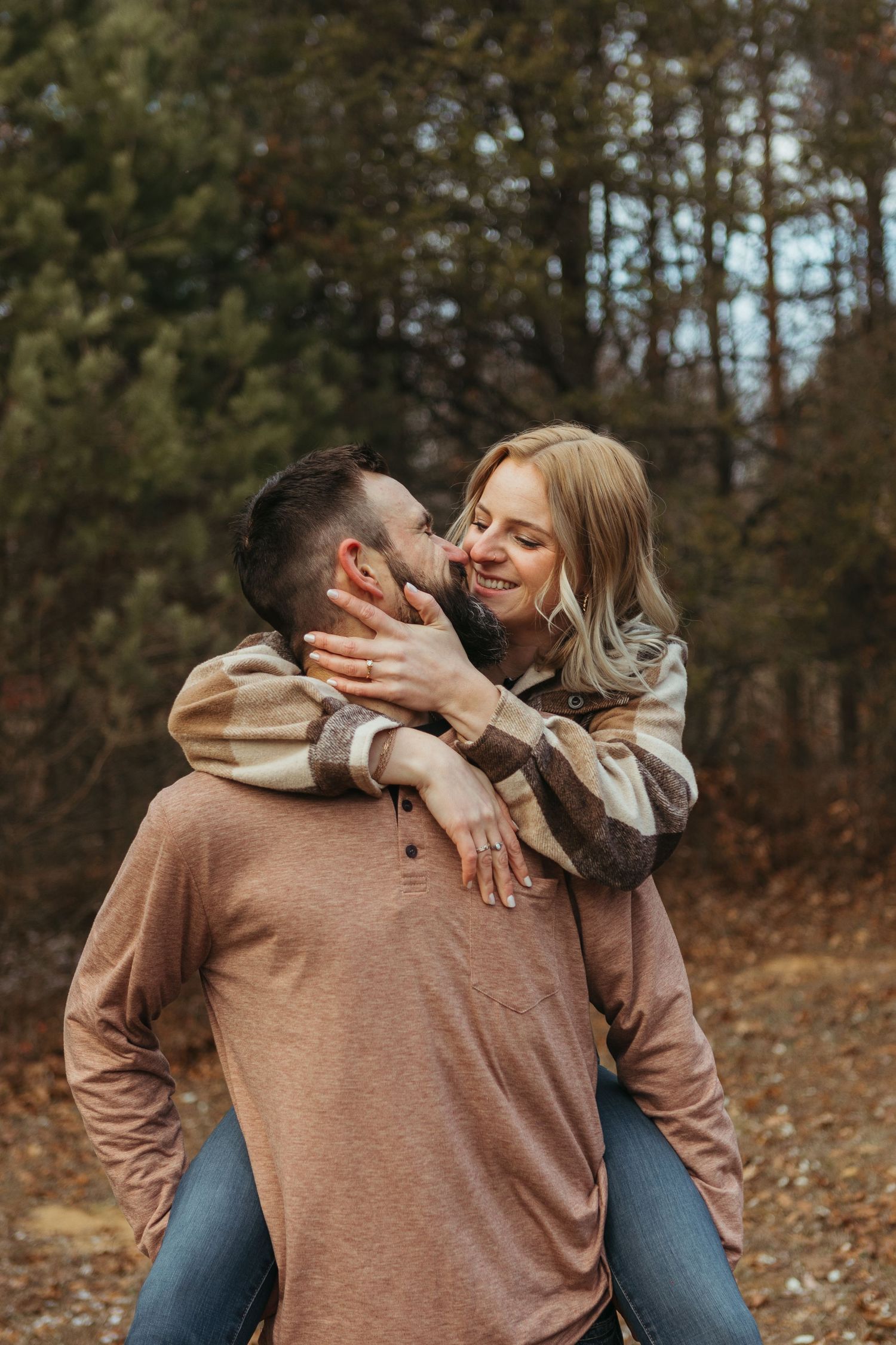 A woman playfully kisses a bearded man giving her a piggyback ride in an autumn forest setting.