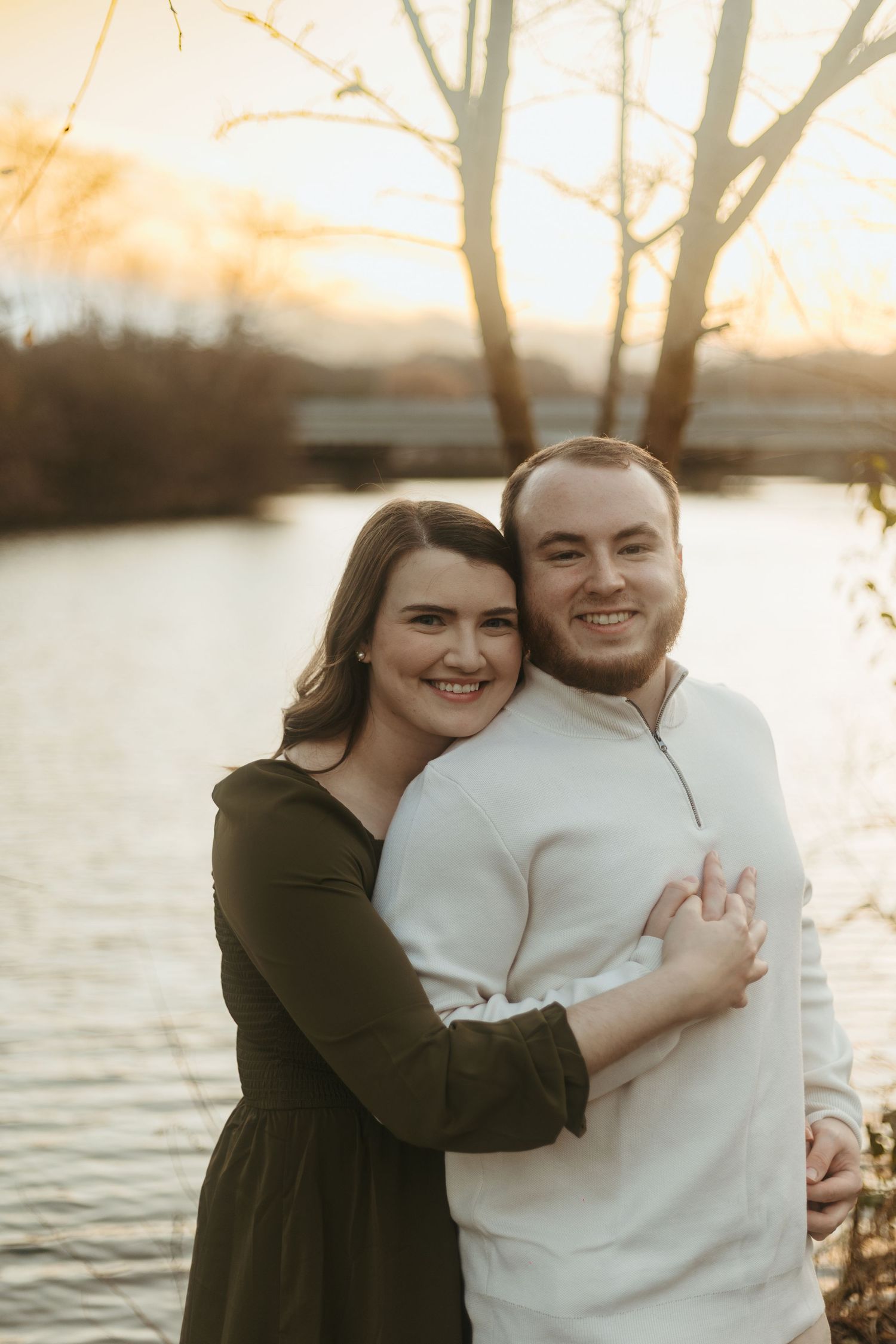 Smiling couple hugging by river at golden sunset near bare trees.
