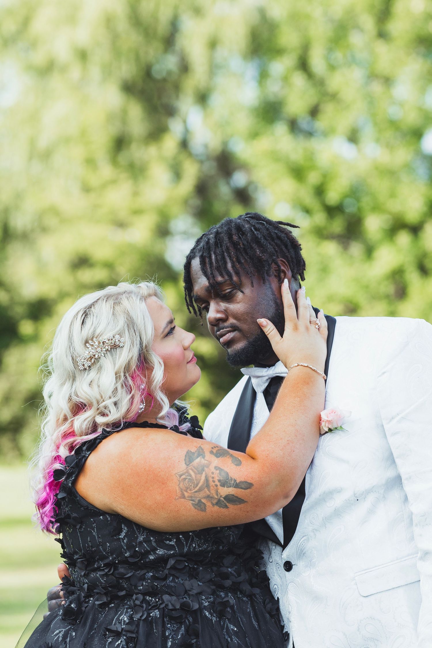 Interracial couple in wedding attire embracing outdoors, bride in black dress with pink hair and rose tattoo.