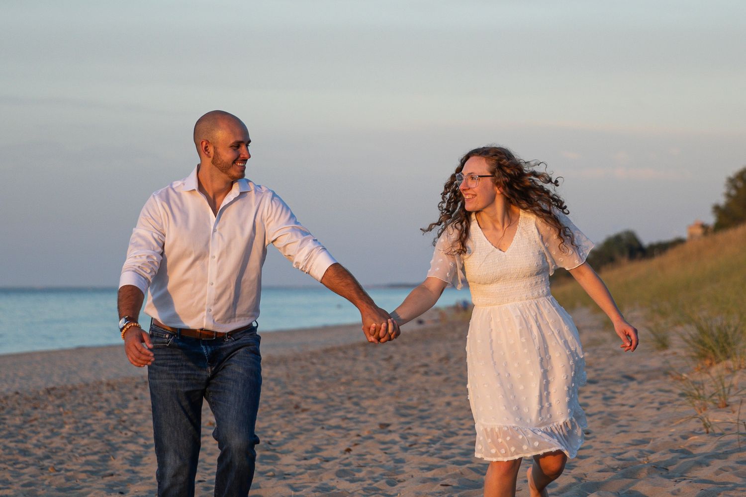 Happy couple walking hand in hand on a sandy beach at sunset, woman in white dress.