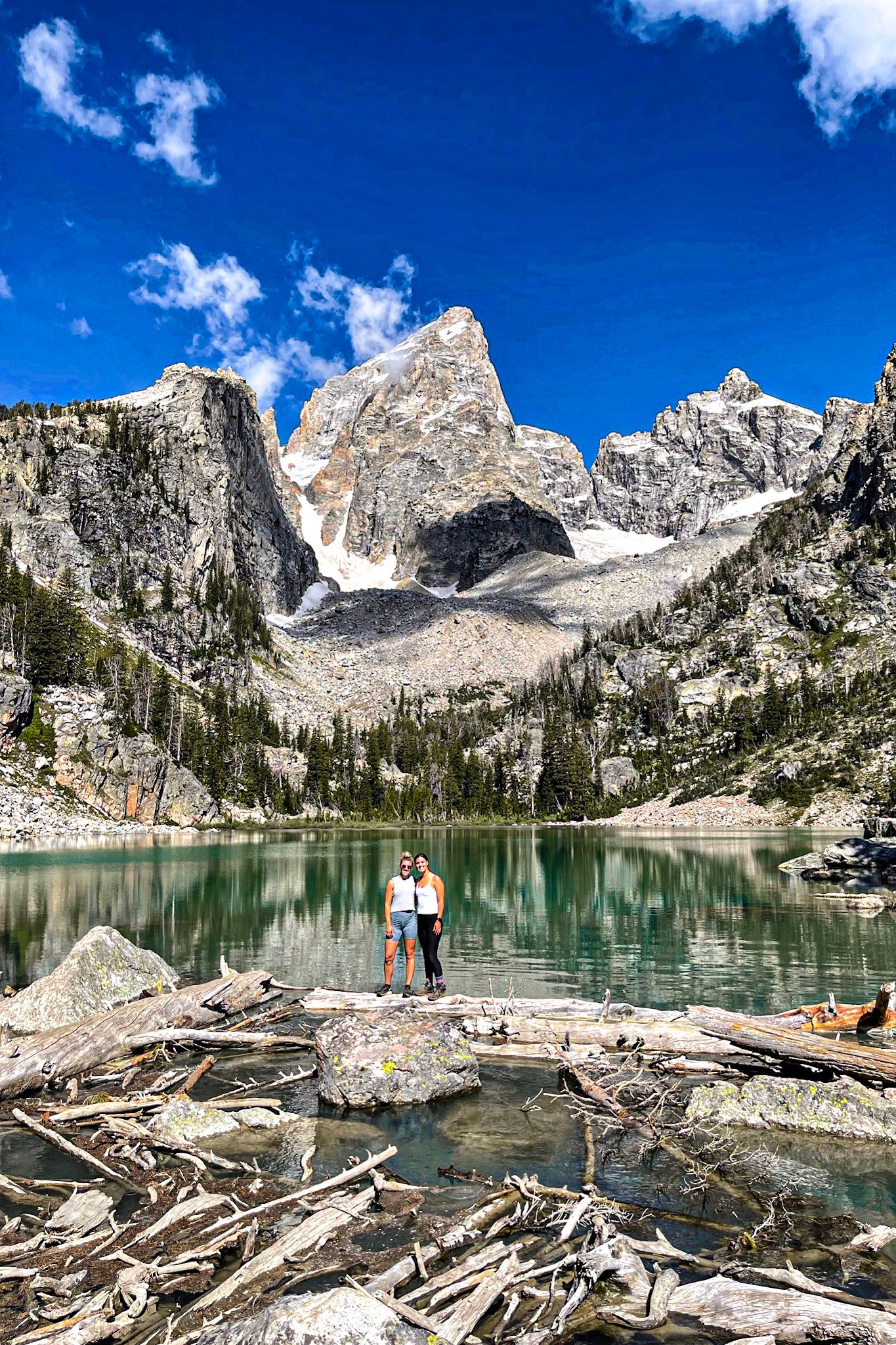 Hikers standing along turquoise Delta Lake beneath towering Grand Teton peaks on a clear summer day.