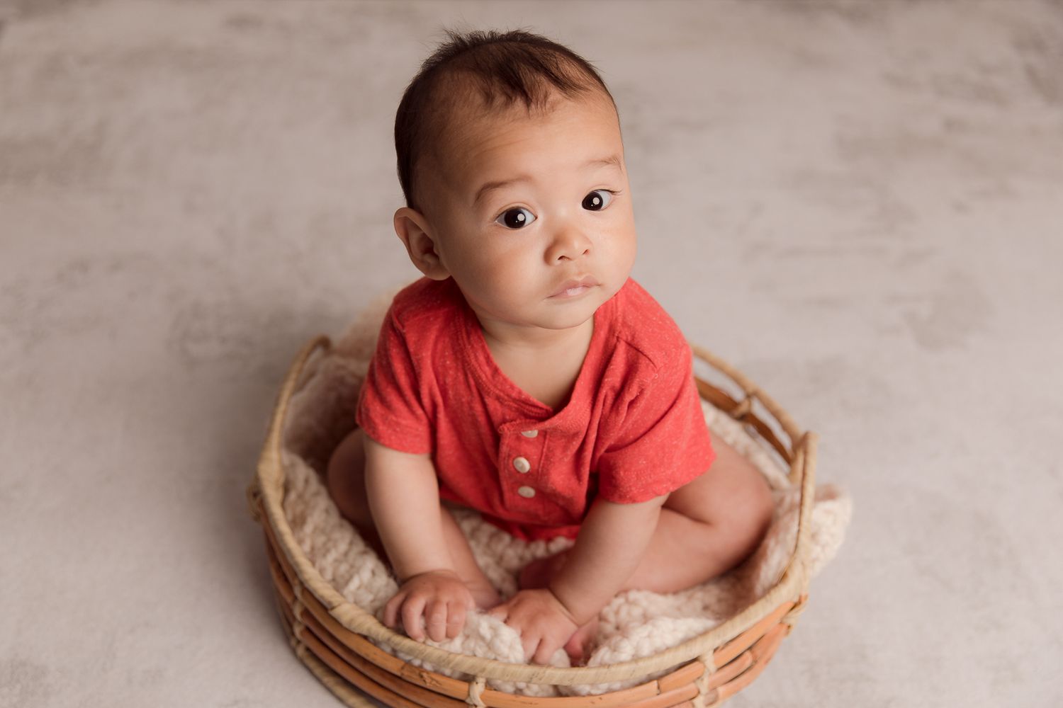 Adorable baby in red onesie sitting in a wicker basket on a gray background. Vista Newborn Photographer