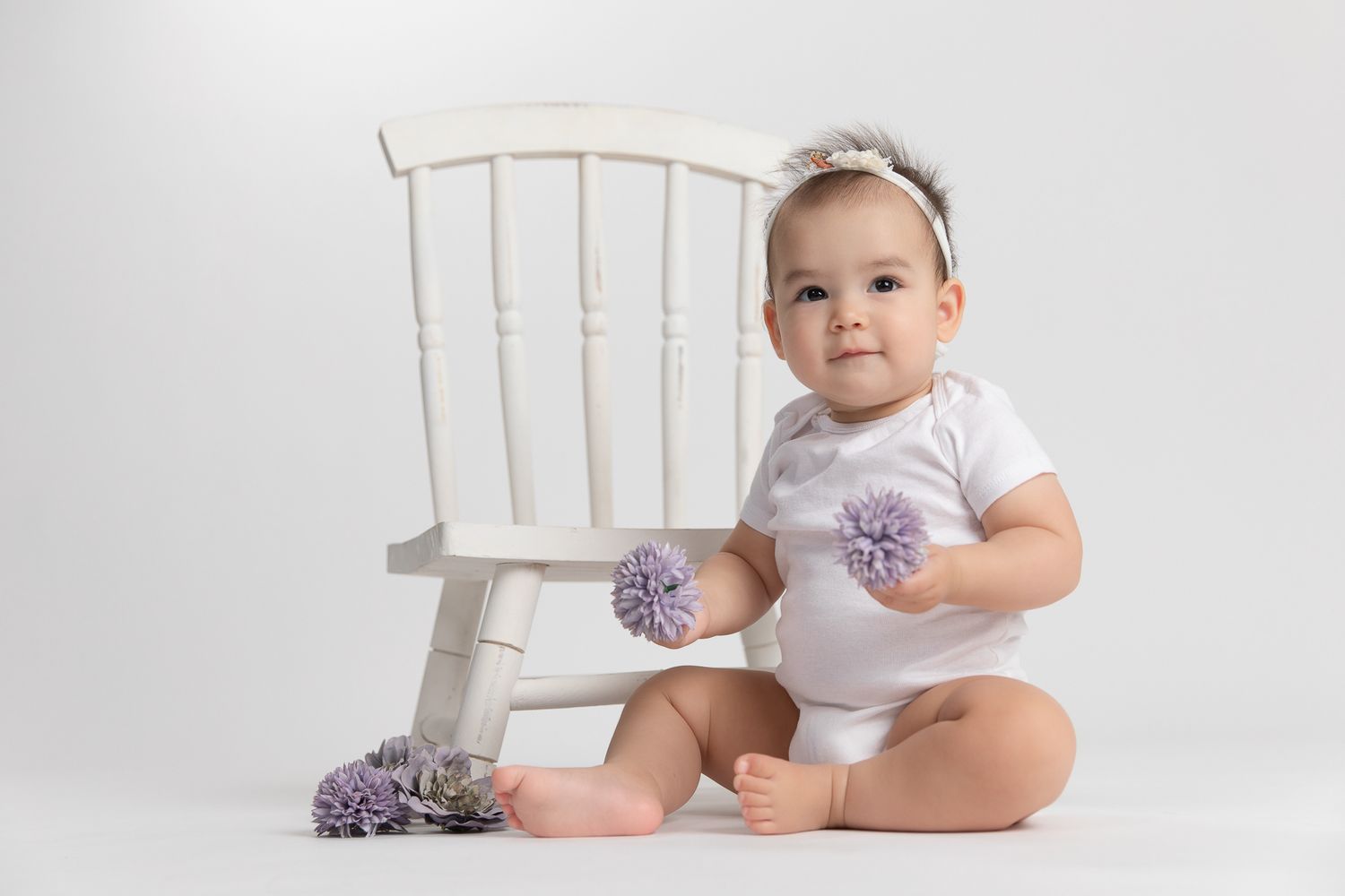 Baby girl in white onesie with purple pom poms sitting beside white chair.