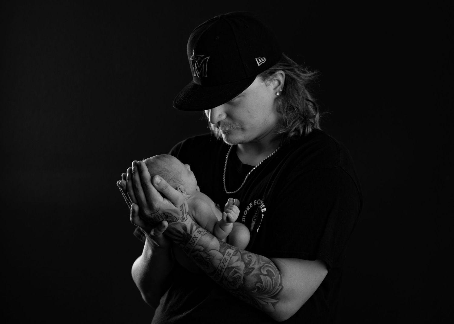Black and white portrait of tattooed person in cap holding newborn baby, studio photography.