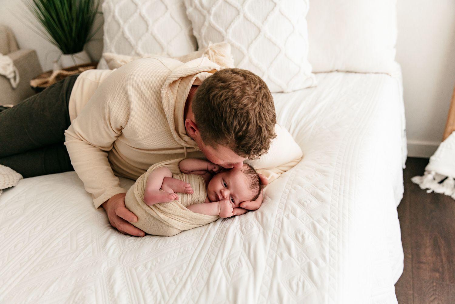 A father tenderly kisses his swaddled newborn baby on a white bed.