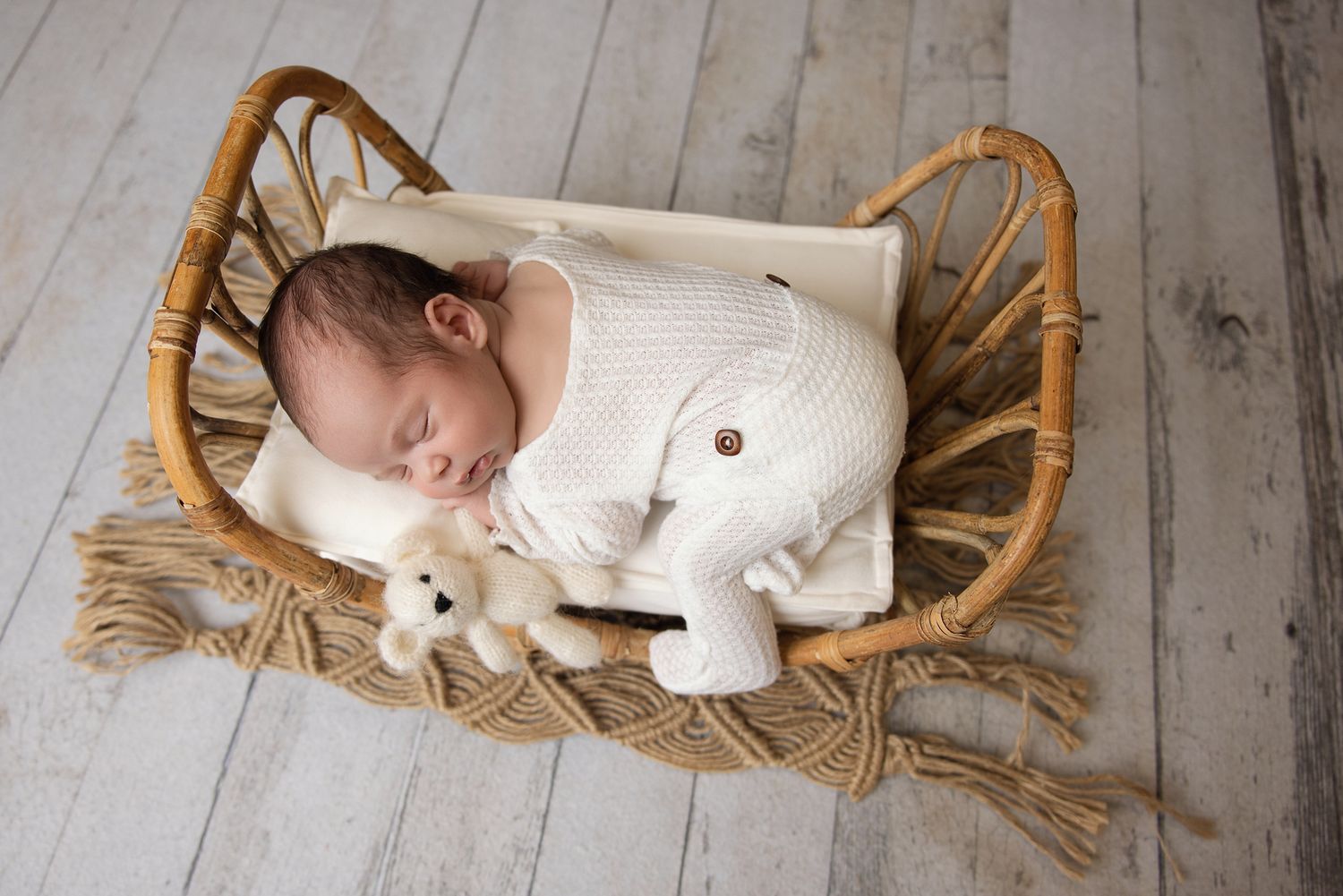 Newborn baby sleeping peacefully in a wicker bassinet wearing a cream knit outfit, holding a small teddy bear.