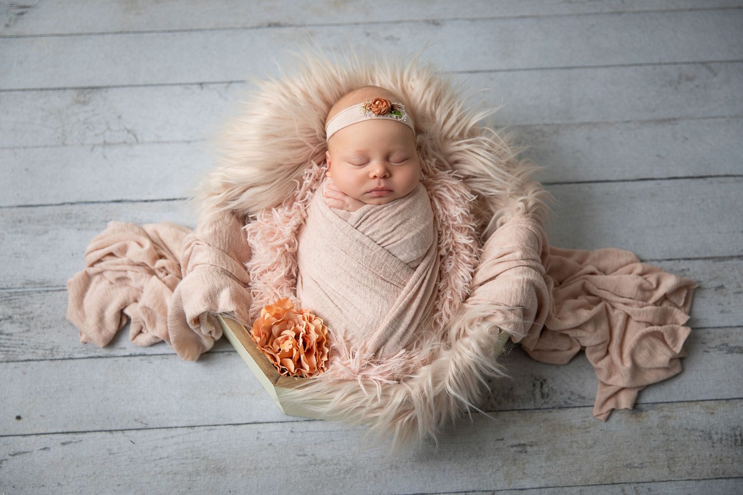 Newborn baby girl swaddled in pink wrap sleeping in a fur-lined basket with floral headband on wood floor.