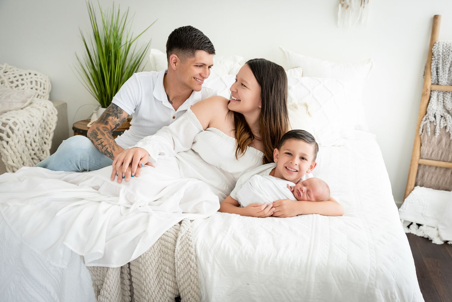 Family of four with newborn baby relaxing together on a white bed in a bright, cozy bedroom setting.