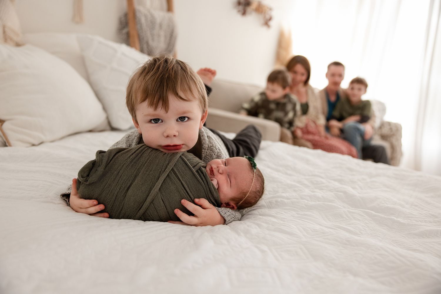Young boy lying on white bed holding swaddled newborn baby, family with parents and siblings sitting in background.