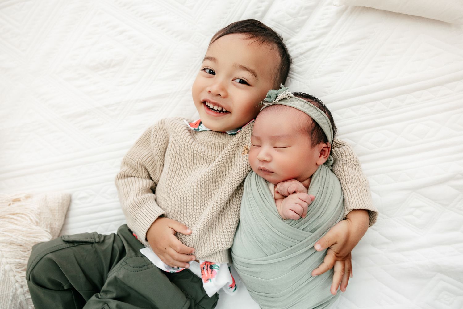 Smiling toddler boy lying next to sleeping newborn baby wrapped in mint green swaddle on white bed.
