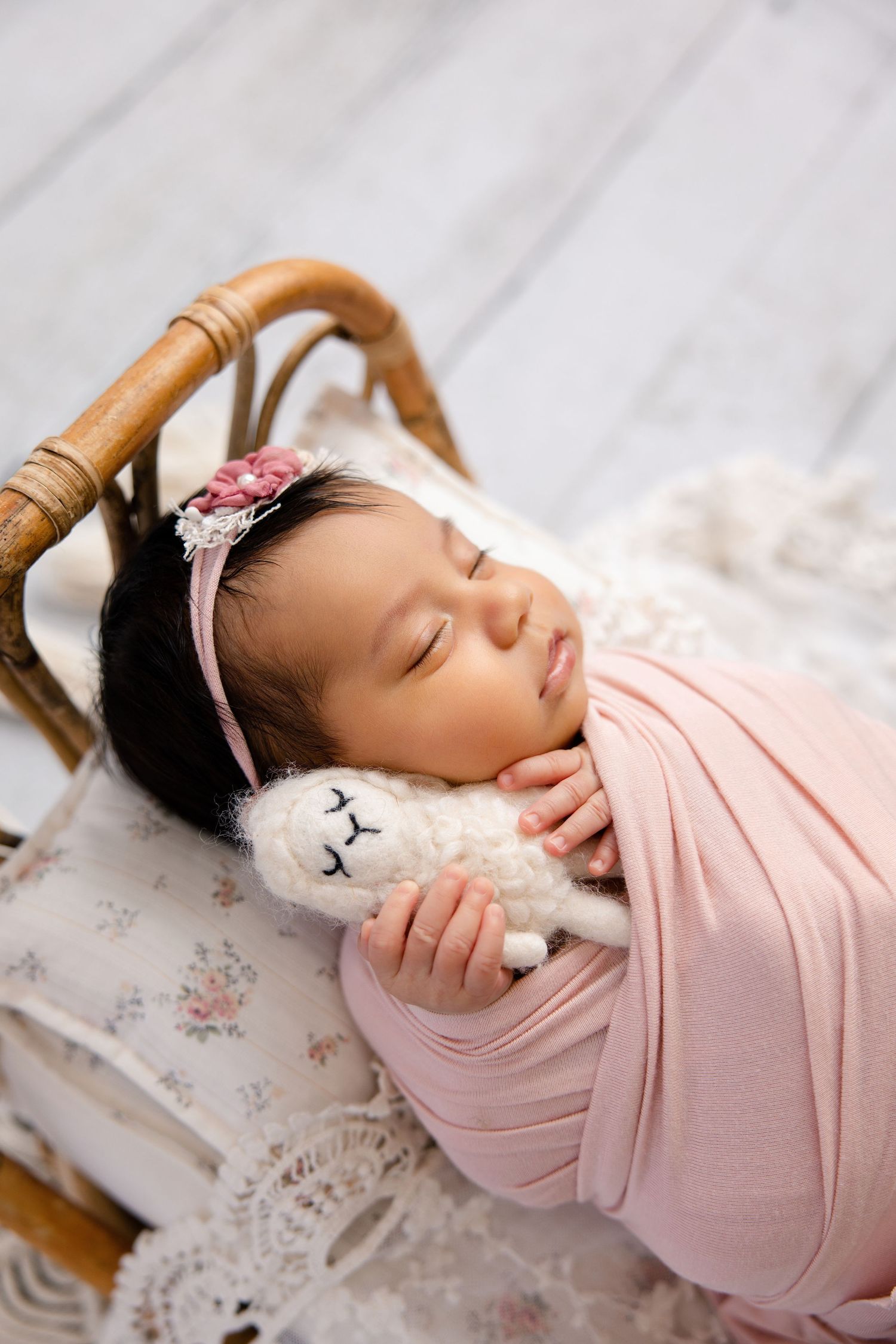 Sleeping newborn baby girl wrapped in pink swaddle holding a stuffed lamb toy in a wicker basket.