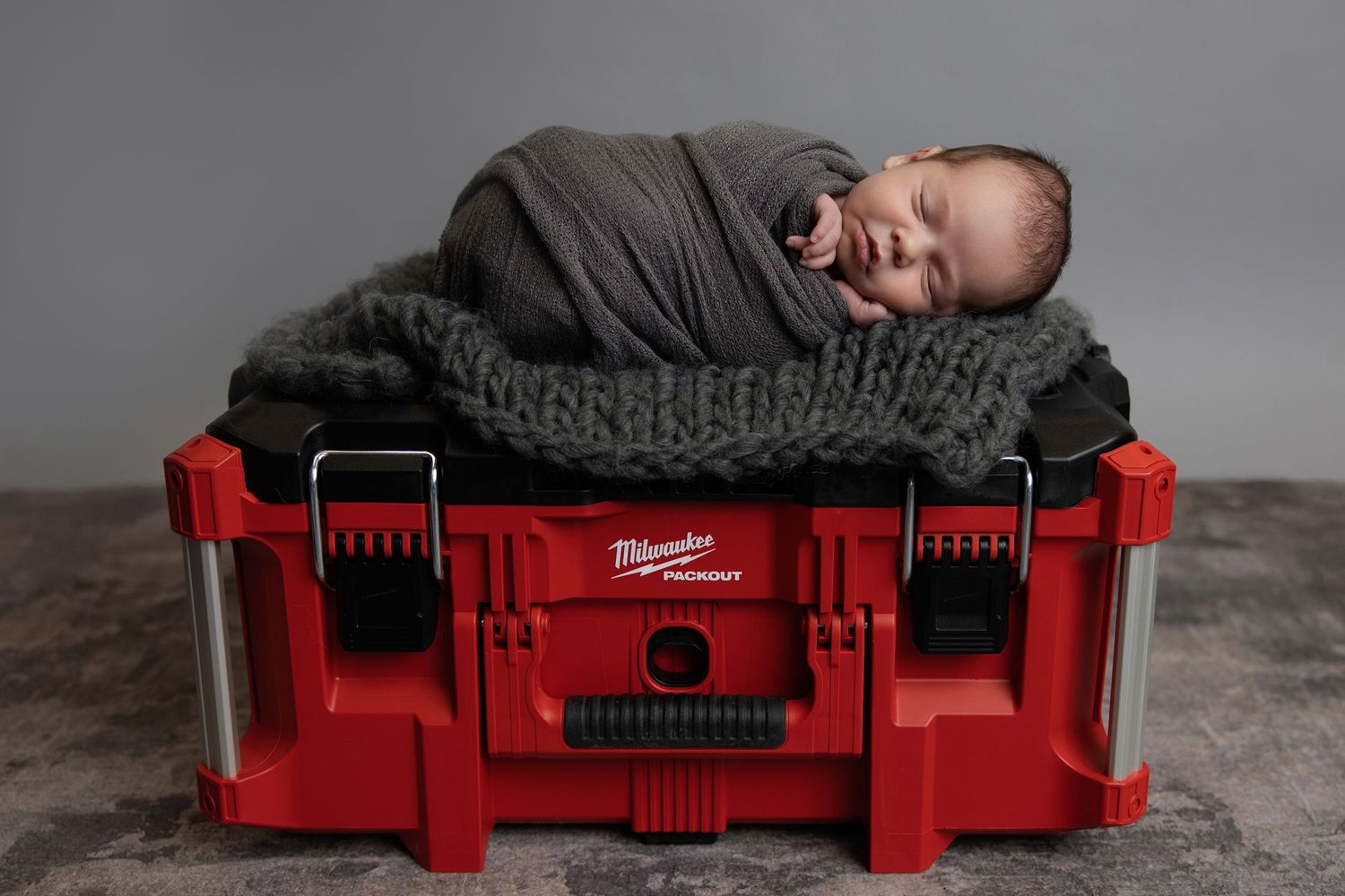 Smiling newborn baby wrapped in gray swaddle sleeping on knit blanket atop a red Milwaukee Packout tool case.