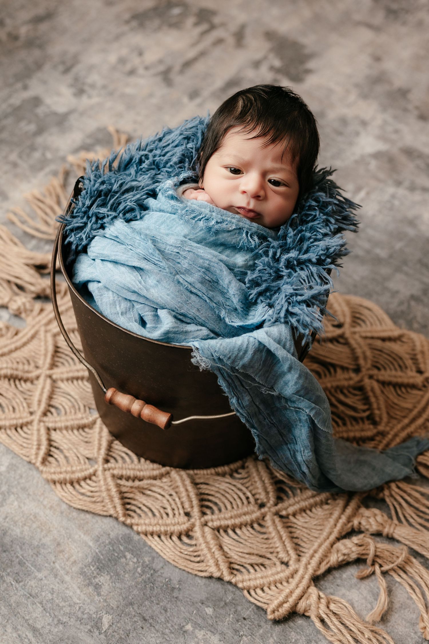 Newborn baby wrapped in blue fringe cloth inside a dark bucket on a woven macrame mat.
