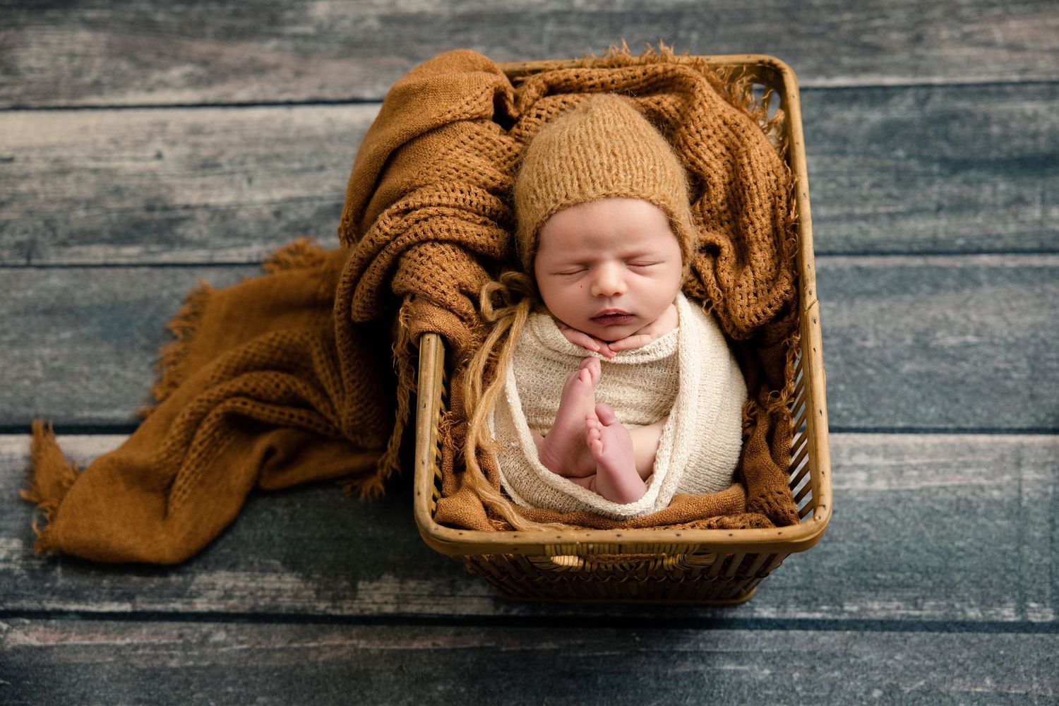 Newborn baby sleeping peacefully in a wicker basket with a golden knit blanket and bonnet on a rustic wood floor.