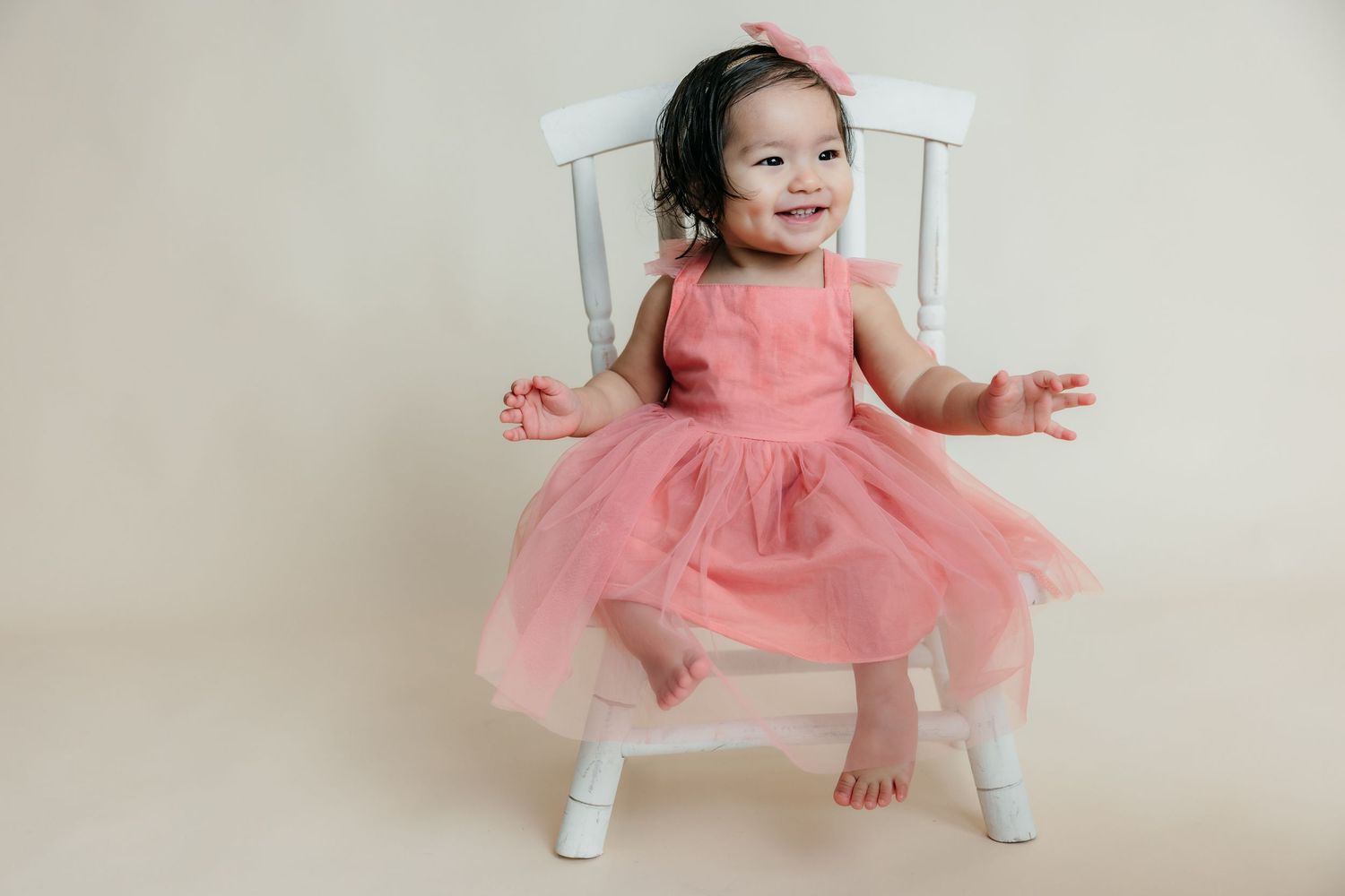 Adorable baby girl in a pink tulle dress sitting on a white chair, smiling happily during her first birthday photoshoot.