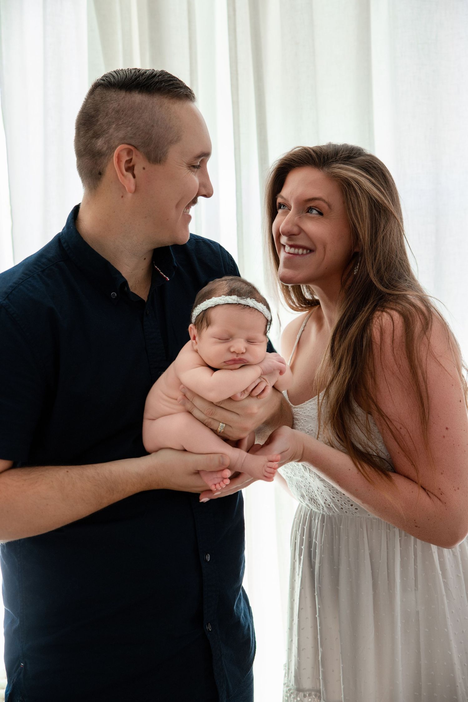 Happy parents smiling at their sleeping newborn baby with a headband during a professional newborn photography session.