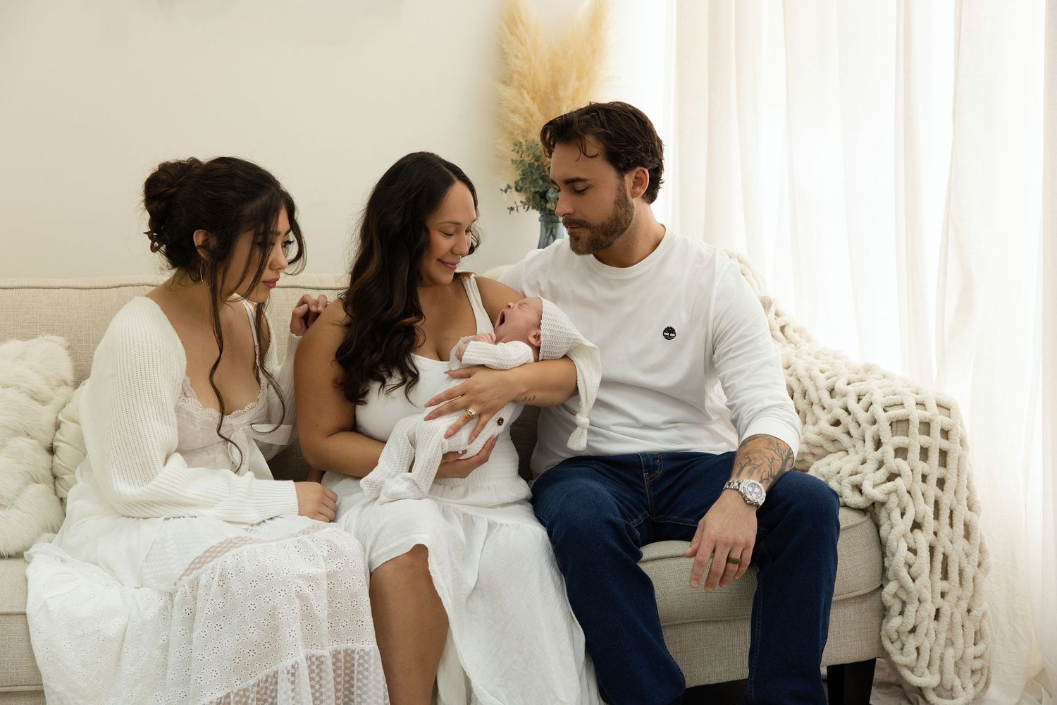 Family of three with newborn baby sitting on a couch, parents smiling at infant in bright white room.