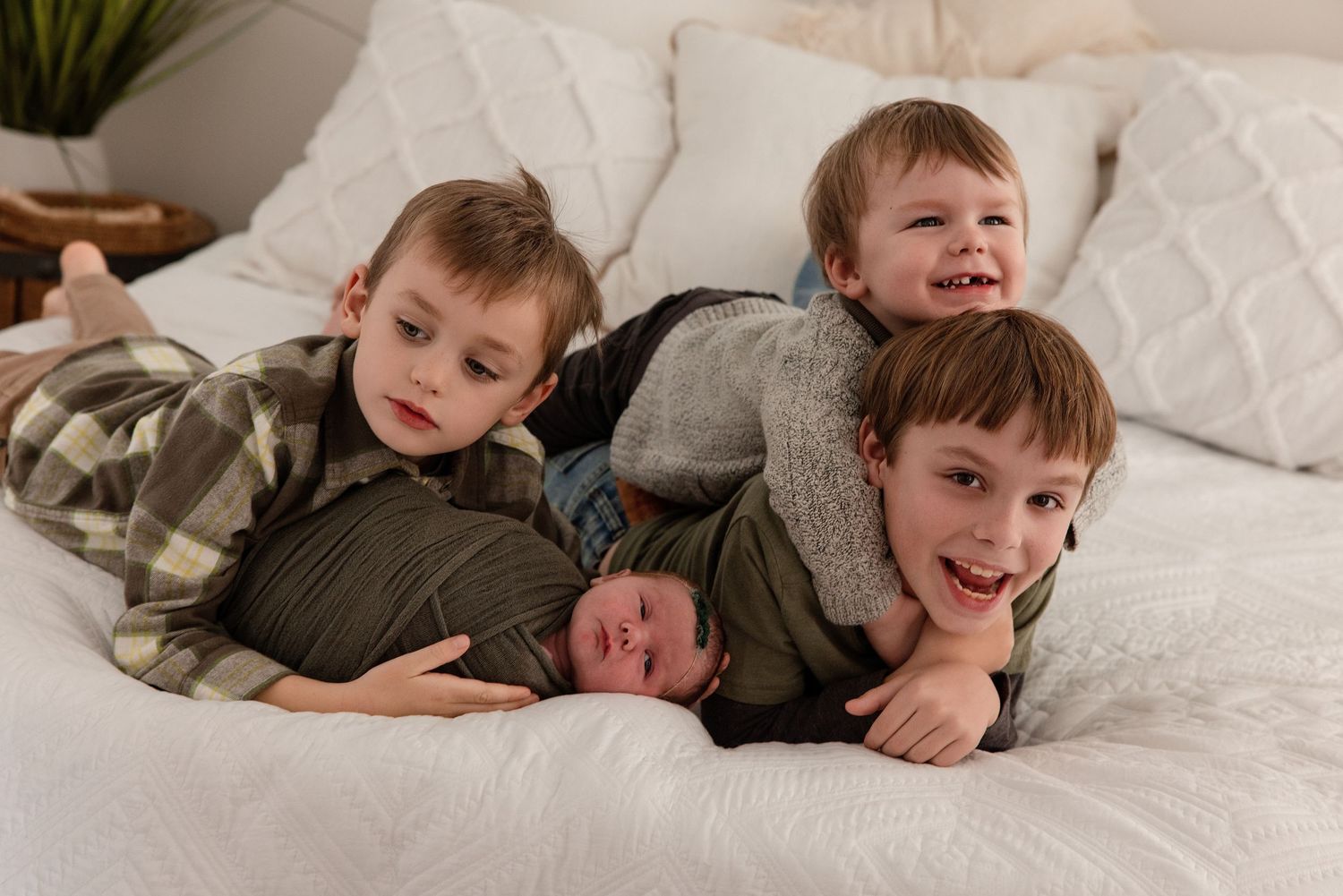 Three young brothers pile on a bed smiling, cradling their newborn sibling in a cozy family portrait.