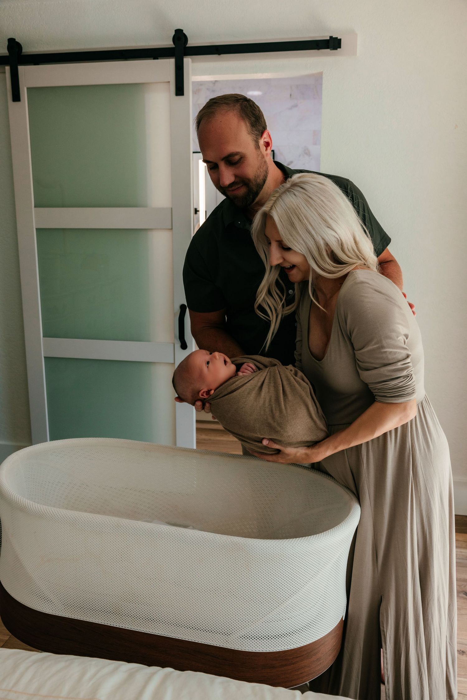 New parents stand together over a bassinet, tenderly holding their swaddled newborn baby.
