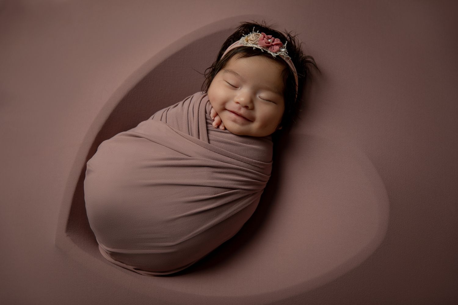 Smiling swaddled newborn baby girl in a floral headband poses in a mauve heart-shaped backdrop. vista newborn photographer
