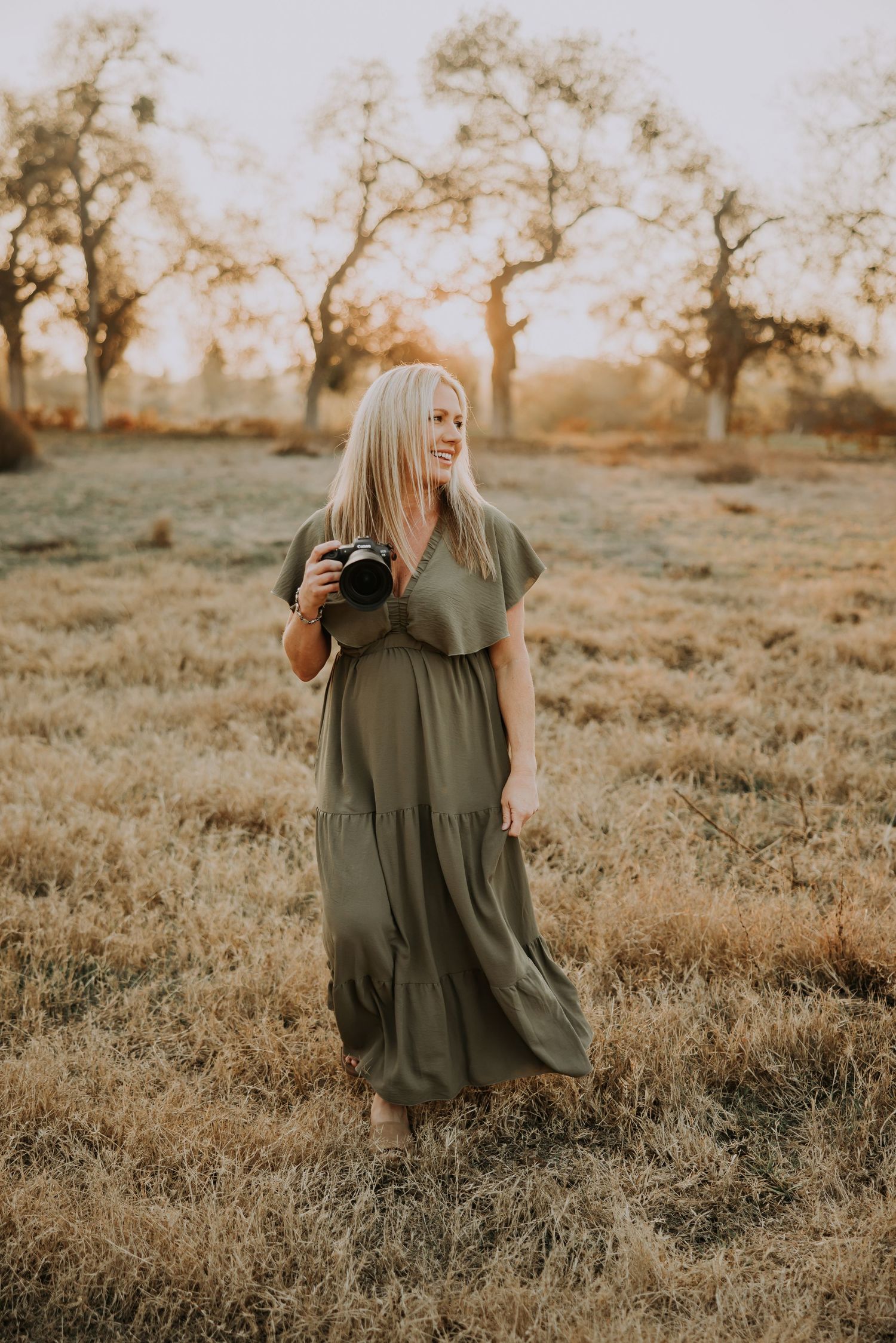 A photographer in a flowing olive green dress stands in a golden field at sunset with trees in the background.