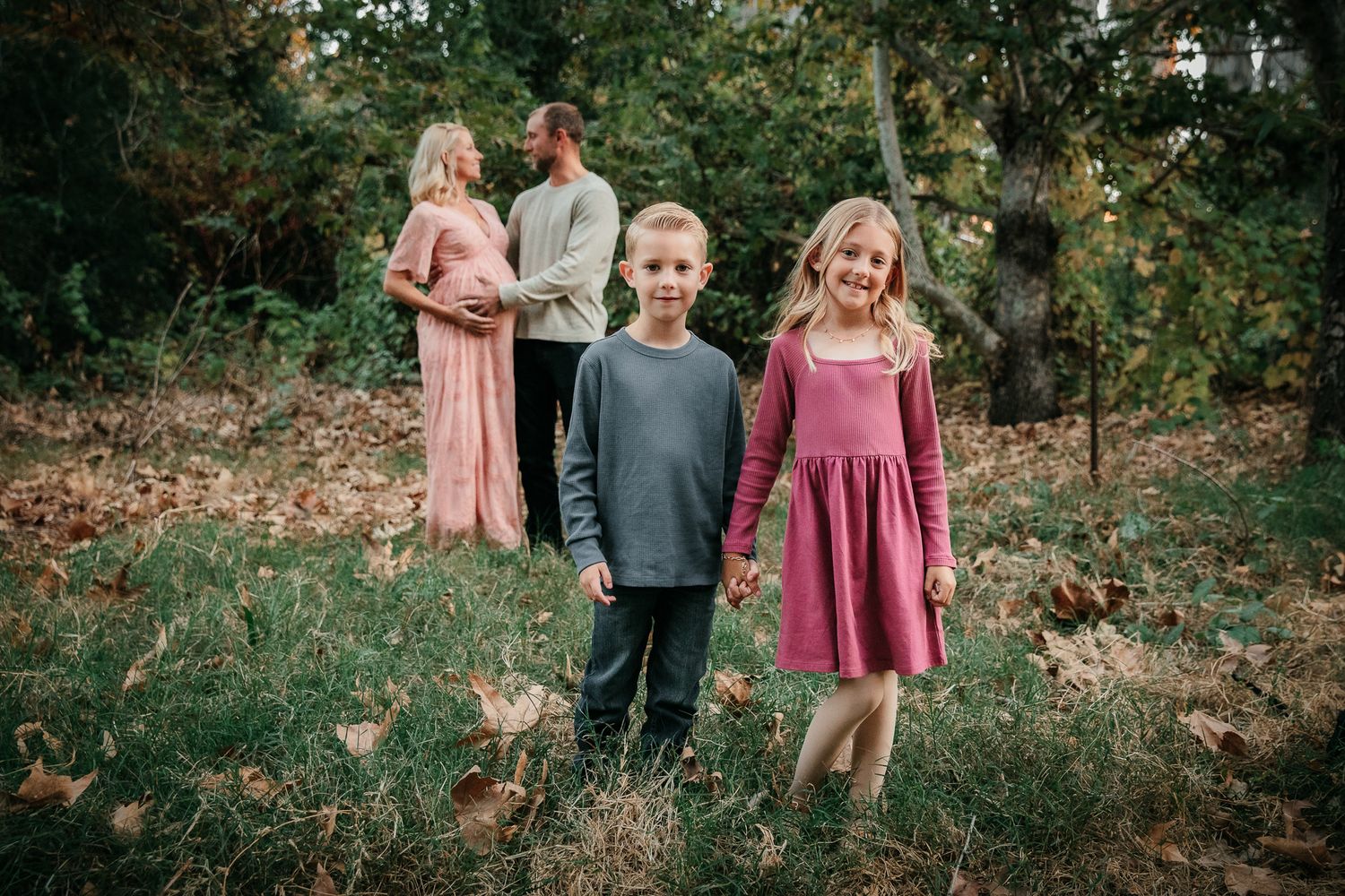 Two siblings holding hands in foreground with pregnant parents embracing in autumn woods.  In Bonsall, near Oceanside