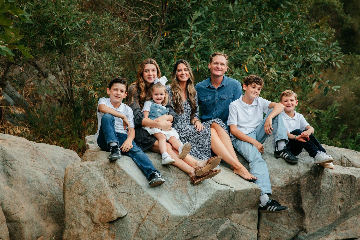 Large family of seven sitting on rocks outdoors surrounded by lush green trees in a casual portrait in Fallbrook, CA