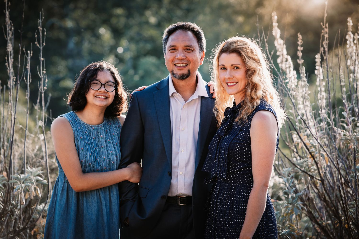 A trio of two women and a man smile together outdoors in golden sunlight among tall grasses.