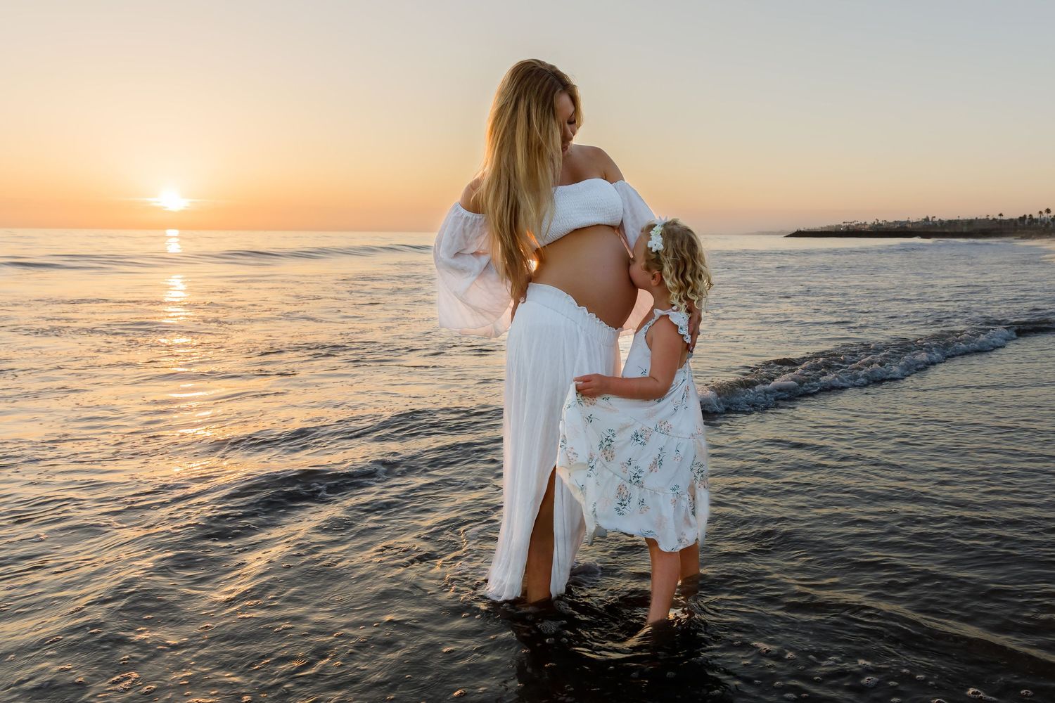 Pregnant mother in white outfit embracing young daughter at sunset on a serene beach.
