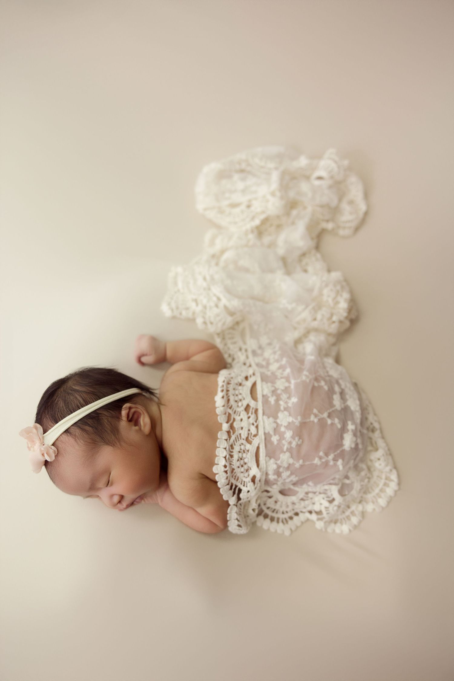 Newborn baby girl sleeping peacefully wrapped in white lace fabric with a delicate pink flower headband.