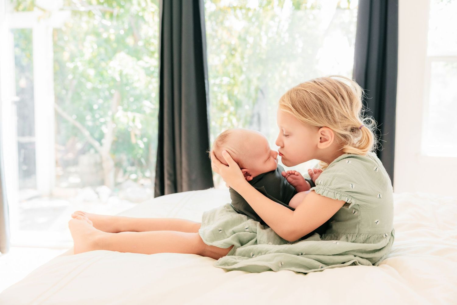 Young blonde girl sitting on white bed kissing newborn baby sibling near bright window with green curtains.