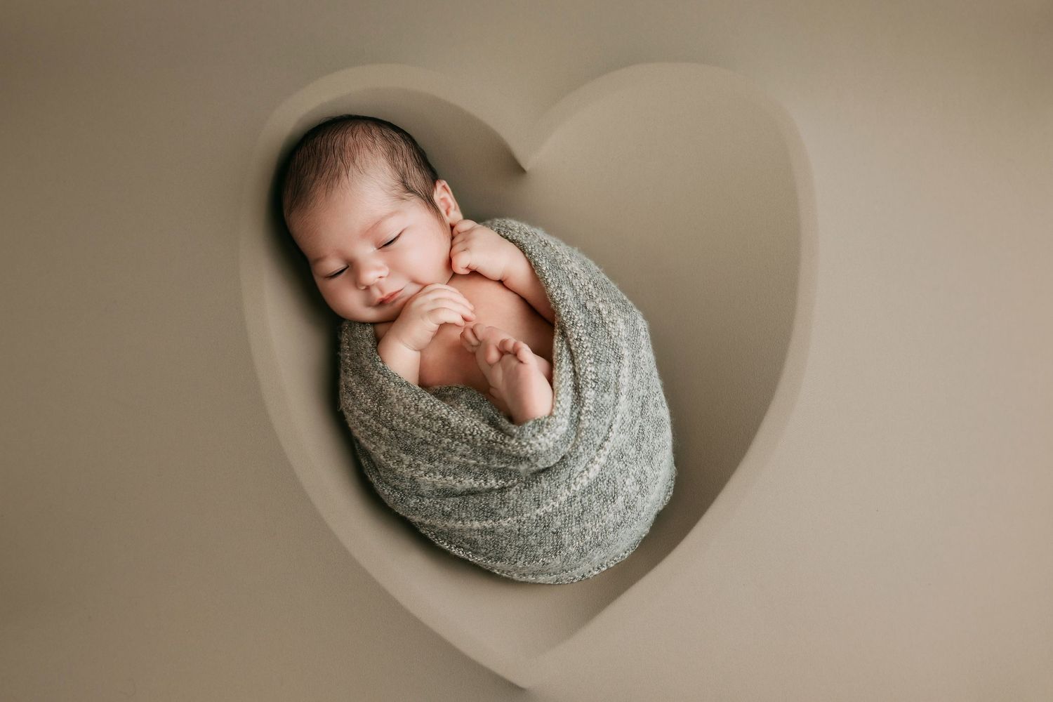 Peaceful newborn baby swaddled in sage green wrap sleeping inside a heart-shaped cutout prop.