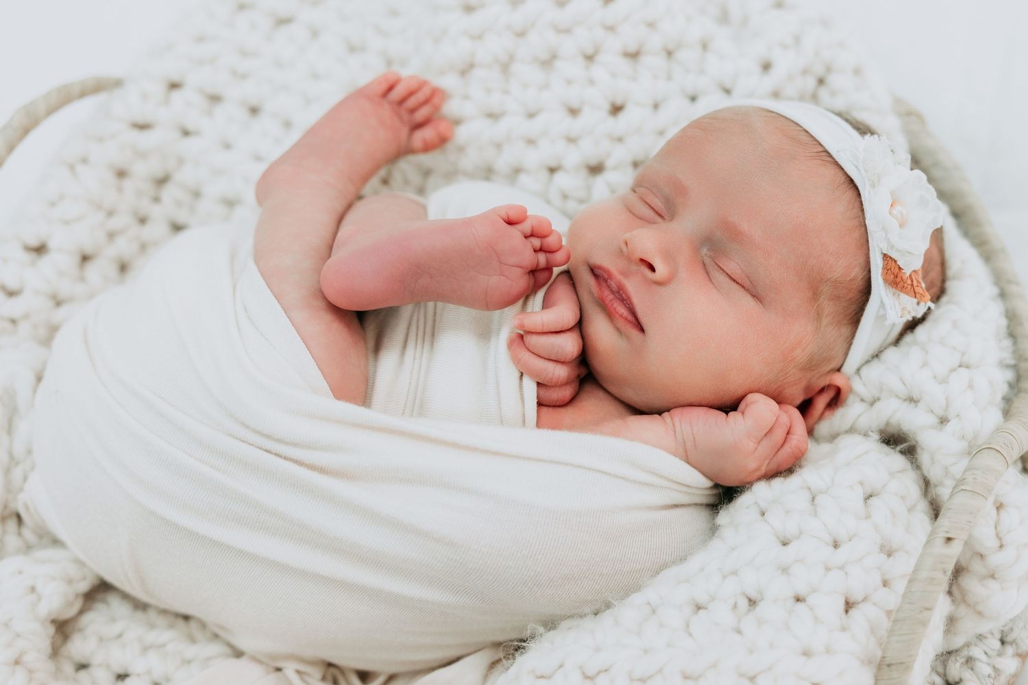 Peaceful newborn baby swaddled in white wrap sleeping in a cozy knit basket with a small butterfly clip.